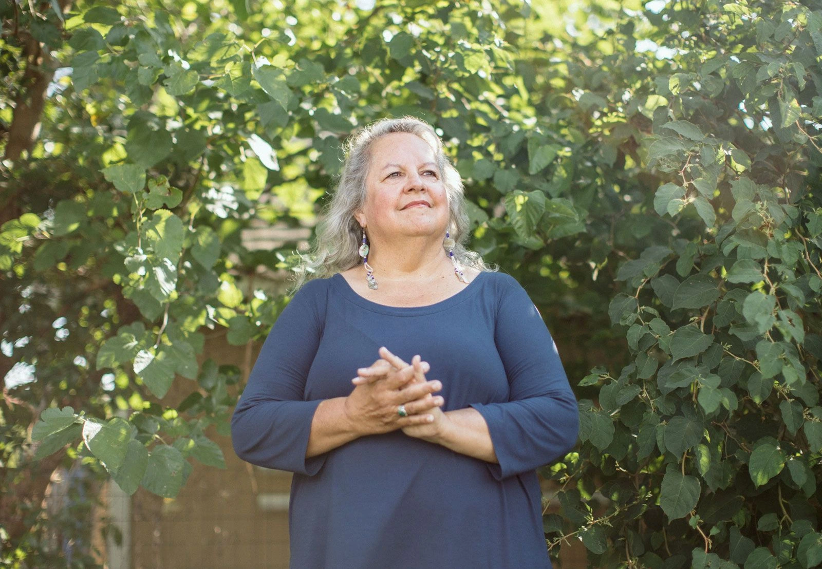Robin Wall Kimmerer staat buiten tussen groen, met gevouwen handen en een rustige, documentaire uitstraling.